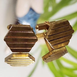 May include: A pair of gold-toned cufflinks with a geometric, hexagonal design. Each cufflink features horizontal, parallel lines across its face. The cufflinks are displayed on a reflective surface, showcasing their mirrored image.