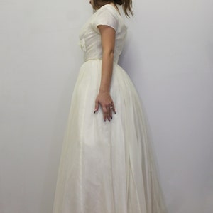 May include: A white vintage wedding dress with a fitted bodice and a full skirt. The dress has a scoop neckline and short sleeves.