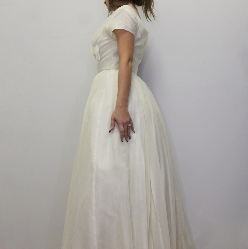 May include: A white vintage wedding dress with a fitted bodice and a full skirt. The dress has a scoop neckline and short sleeves.