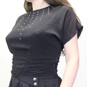 May include: A black, short-sleeved top with a fitted bodice and a gathered waist. The top features a V-shaped neckline adorned with silver rhinestones.