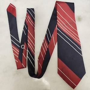 May include: A necktie featuring a diagonal striped pattern in deep navy blue and vibrant red, accented by thin, crisp white lines running parallel across the fabric. The tie displays a classic pointed end and a smooth, woven texture throughout.