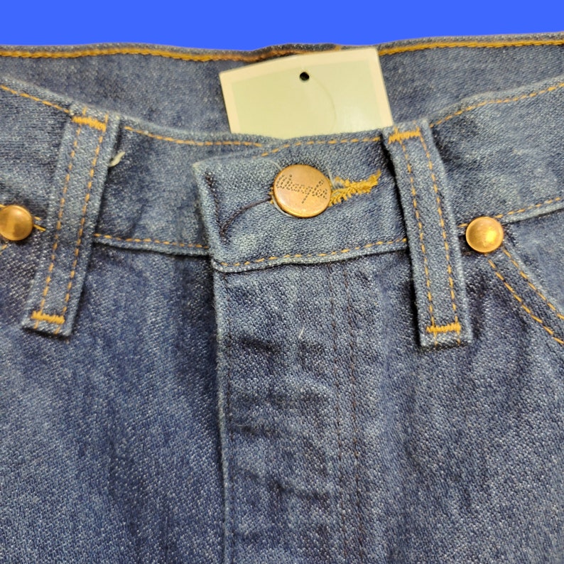 May include: Close-up of a pair of blue denim jeans with a brown leather label that says "Wrangler" on the waistband.