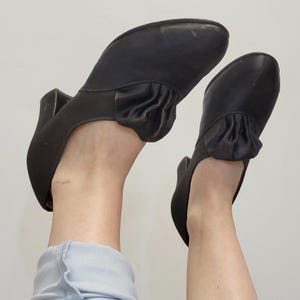 May include: A pair of black vintage heels with a rounded toe and a small heel. The shoes have a decorative ruffle detail on the top of the foot. The shoes are made of a smooth material, possibly leather.