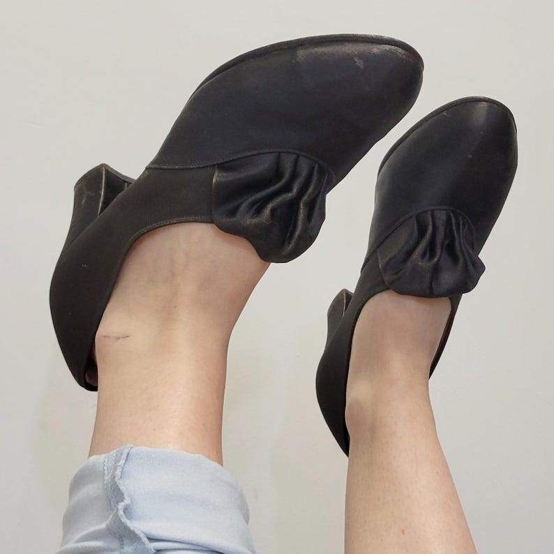 May include: A pair of black vintage heels with a rounded toe and a small heel. The shoes have a decorative ruffle detail on the top of the foot. The shoes are made of a smooth material, possibly leather.