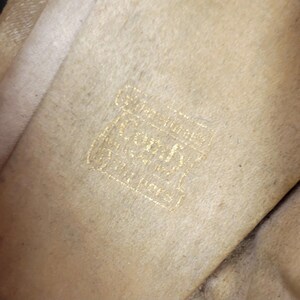 May include: Close-up of a beige fabric with a gold logo. The logo includes the word "Coach" in a decorative font, accompanied by other text. The fabric has a soft texture and a slightly aged appearance, with minor colour variations.