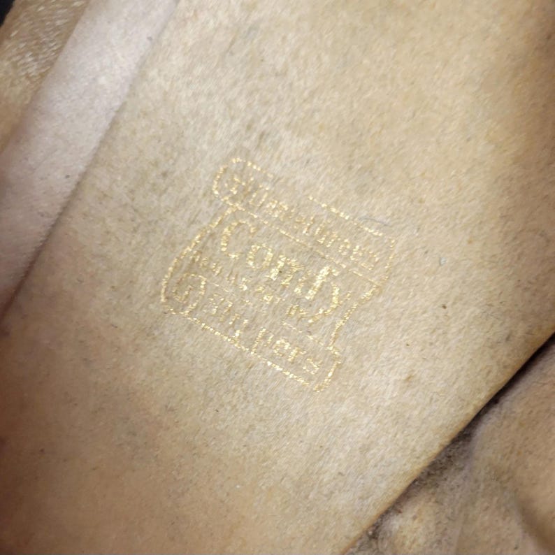 May include: Close-up of a beige fabric with a gold logo. The logo includes the word "Coach" in a decorative font, accompanied by other text. The fabric has a soft texture and a slightly aged appearance, with minor colour variations.