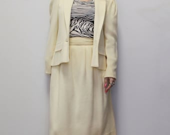May include: A cream-coloured skirt suit with a blazer and a knee-length skirt. The blazer has a notched collar and long sleeves. The skirt has a high waist and a straight silhouette.