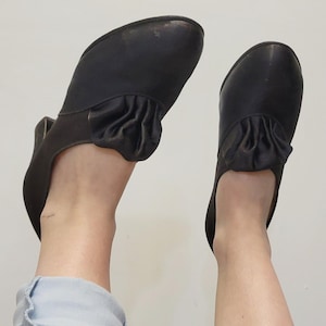 May include: A pair of black shoes with a rounded toe and a small heel. The shoes feature a gathered fabric detail on the top. The shoes are being worn. The shoes are black.
