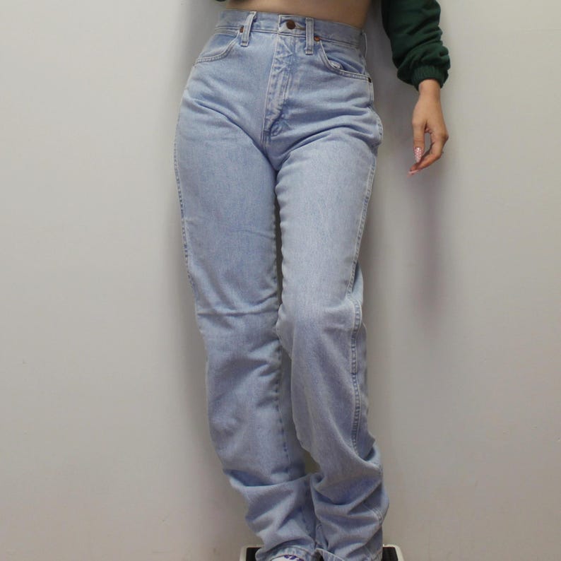May include: High-waisted light wash denim jeans featuring a classic five-pocket design, a metal button closure, and a zip fly. These straight-leg jeans have a faded blue finish with visible stitching along the seams and a structured waistband.