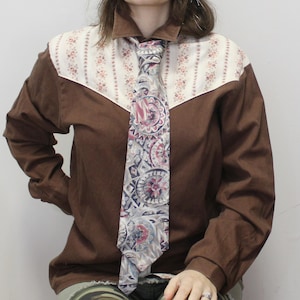 May include: A brown long-sleeved shirt with a cream-colored yoke featuring a floral pattern. A patterned tie in shades of gray, pink, and blue is knotted at the collar. The shirt has a classic western style.