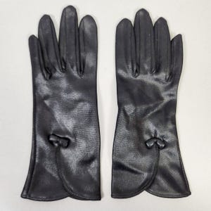 May include: A pair of black gloves. The gloves are made of a smooth material and have a small bow detail on the back of the wrist. The gloves are long, extending past the wrist.