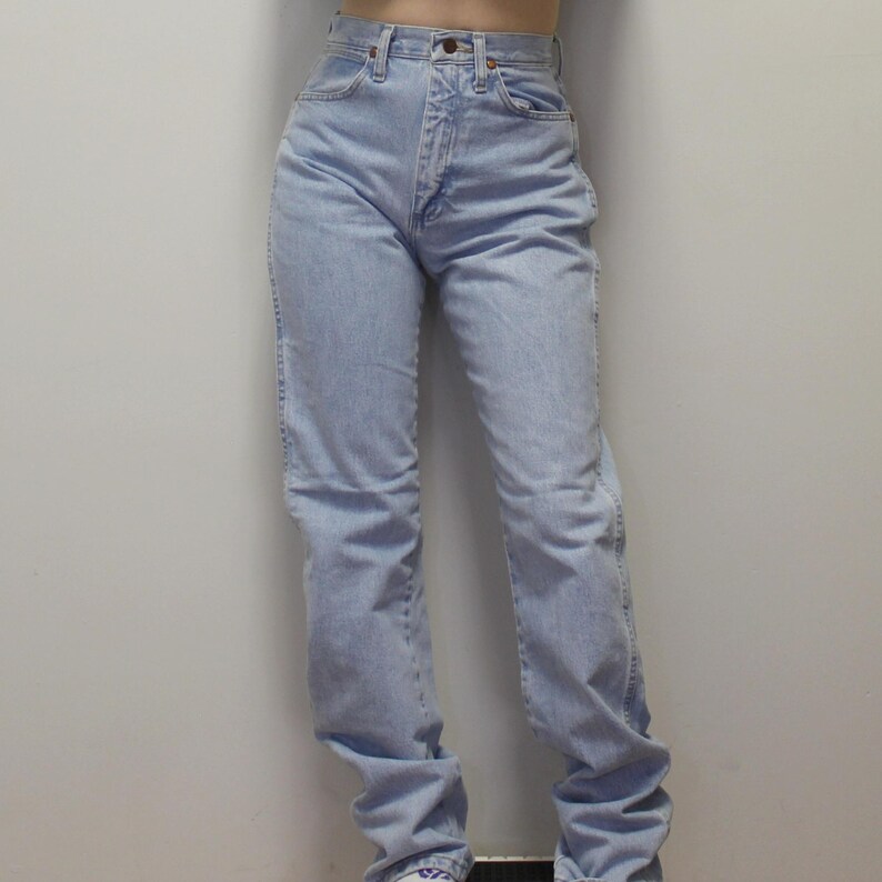 May include: High-waisted light blue denim jeans featuring a classic five-pocket design, a metal button closure, and a zip fly. The jeans have a straight-leg cut with visible stitching along the seams and a light wash finish throughout the fabric.