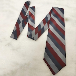 May include: A necktie featuring a repeating diagonal striped pattern in shades of dark red, slate gray, and silver. The fabric has a smooth, satin-like finish with a pointed tip at the bottom and a folded section showing the reverse side.