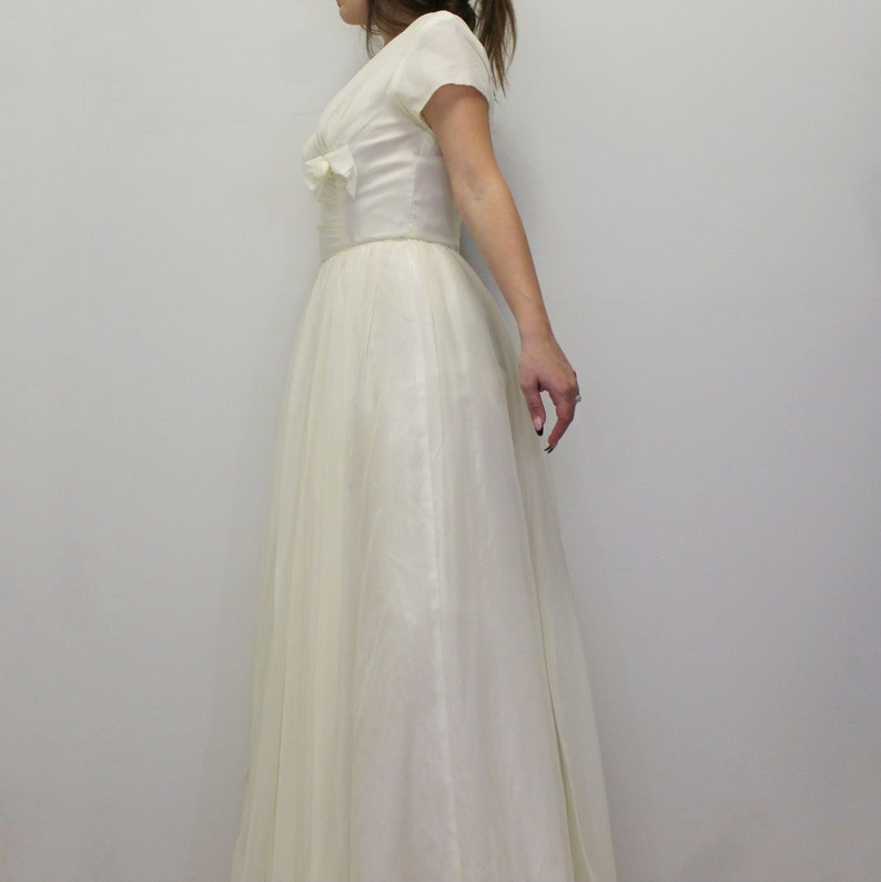 May include: A white vintage wedding dress with a fitted bodice and a full skirt. The dress has a V-neckline and short sleeves.