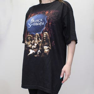 May include: Black t-shirt with a graphic of the band Black Sabbath. The graphic features the band members in a forest setting with a blue circle behind them that says "Black Sabbath".