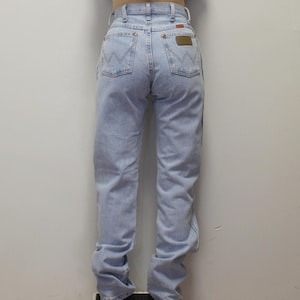 May include: Light blue denim jeans featuring a high-waisted fit, belt loops, and two back pockets with a signature W-shaped stitch pattern. A small rectangular brown leather patch is visible on the right back pocket of these classic denim pants.