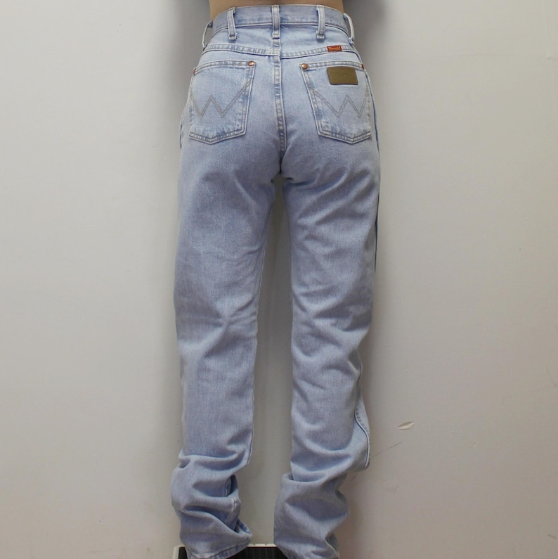 May include: Light blue denim jeans featuring a high-waisted fit, belt loops, and two back pockets with a signature W-shaped stitch pattern. A small rectangular brown leather patch is visible on the right back pocket of these classic denim pants.