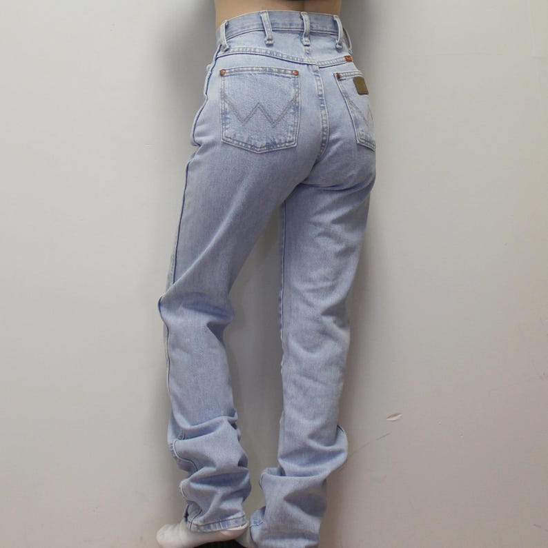 May include: Light blue high-waisted denim jeans featuring a classic five-pocket design, a button and zipper fly closure, belt loops, and a signature W-shaped stitch pattern on the back pockets with a small brown leather brand patch on the rear.
