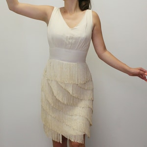 Puede incluir: Vestido sin mangas color crema con cuello en V y cintura entallada. La falda presenta múltiples capas de flecos, creando un aspecto texturizado estilo flapper. El vestido es de color neutro, adecuado para diversas ocasiones.