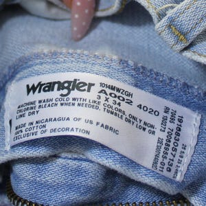 May include: A light blue denim garment interior featuring a white rectangular care label with the Wrangler logo. The label includes text: 1014MWZGH, A002 4020, 3 X 34, 191683057136, 7008955-011, RN 130273, 22E1001440920, 28 5/8 25 3/4NCS, 100% cotton, made in Nicaragua of US fabric, and washing instructions.