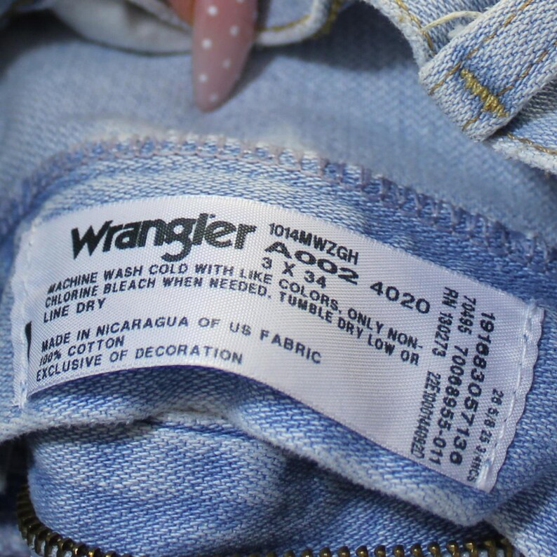 May include: A light blue denim garment interior featuring a white rectangular care label with the Wrangler logo. The label includes text: 1014MWZGH, A002 4020, 3 X 34, 191683057136, 7008955-011, RN 130273, 22E1001440920, 28 5/8 25 3/4NCS, 100% cotton, made in Nicaragua of US fabric, and washing instructions.