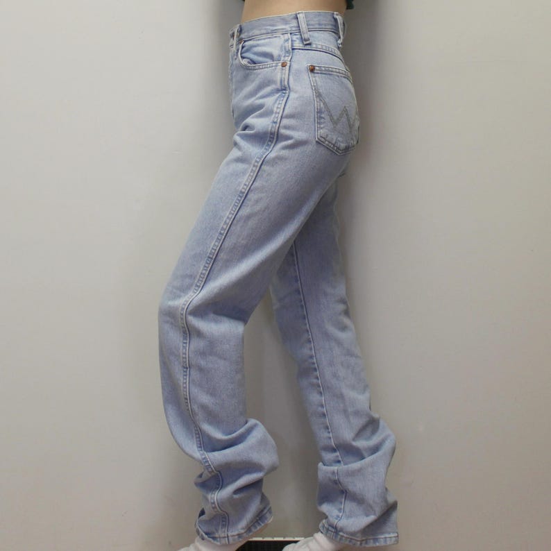 May include: High-waisted light blue denim jeans featuring a straight-leg cut, a button and zipper fly closure, belt loops, and a back pocket with a distinct W-shaped decorative stitch pattern in a lighter thread color on the denim fabric.