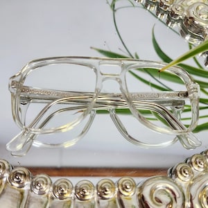 Puede incluir: Gafas transparentes de estilo vintage con doble puente y lentes rectangulares. Las monturas son de plástico transparente con detalles metálicos. Las gafas están sobre una superficie reflectante, con algunas hojas verdes en el fondo. El texto en la montura dice "RAINBOW WEAR USA".