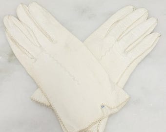 May include: A pair of off-white gloves with a textured surface. The gloves have decorative stitching along the seams and a scalloped edge on the wrist. A small blue tag is visible on one glove.