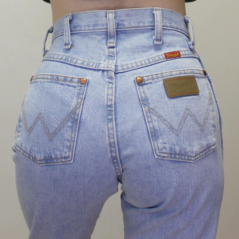 May include: Light blue high-waisted denim jeans featuring a classic five-pocket design, copper-toned rivets, and a brown leather patch on the back pocket with the Wrangler logo. The back pockets display a signature W-shaped decorative stitching.