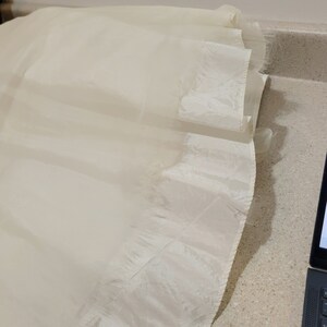 May include: A white tulle fabric with a ruffled edge. The fabric is sheer and has a delicate texture.