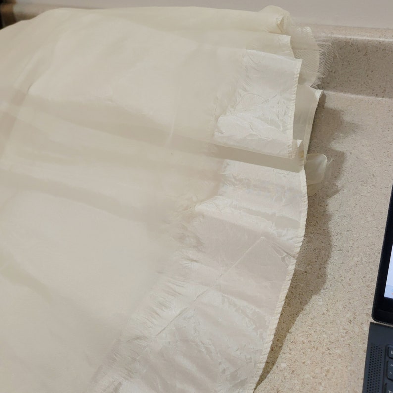 May include: A white tulle fabric with a ruffled edge. The fabric is sheer and has a delicate texture.