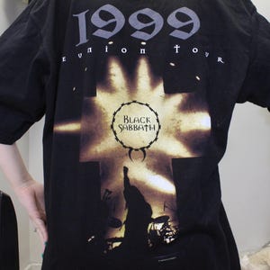 May include: Black t-shirt with a graphic of a cross and a silhouette of a band performing. The text "1999 Reunion Tour" and "Black Sabbath" are printed on the shirt.