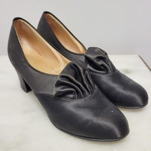 May include: A pair of black satin court shoes with a low heel. The shoes have a gathered fabric detail on the vamp. The interior of the shoe is a light tan colour. The shoes are likely vintage or vintage-inspired.