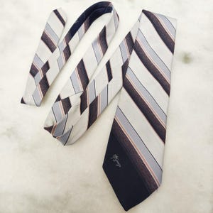 May include: A necktie featuring a diagonal striped pattern in shades of cream, light blue, and dark brown. The wide end of the tie is solid dark brown at the tip and displays the text St. George in a cursive script font. The fabric has a subtle sheen.