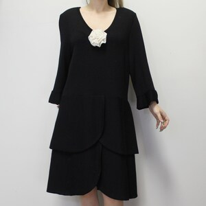 May include: A black dress with a V-neckline and long sleeves. The dress features a white floral accent at the neckline and a layered skirt design. The sleeves have a ruffled cuff.