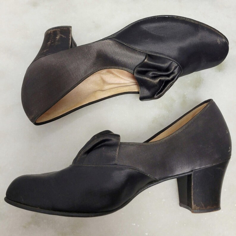 May include: A pair of black satin vintage shoes with a low heel. Each shoe features a decorative bow on the vamp. The shoes have a rounded toe and a sturdy heel.