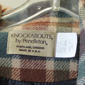 Op de afbeelding: Detail van een label van een geruite wollen jas. Het label luidt "KNOCKABOUTS, by Pendleton, PORTLAND, OREGON MADE IN U.S.A." Een kleiner label geeft maat L en 100% wol aan. De jas is in tinten bruin, groen en blauw.