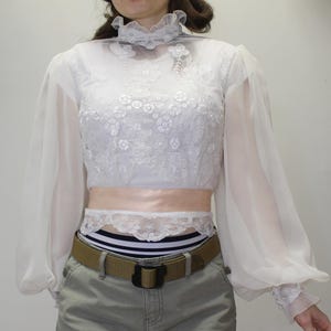 May include: White, long-sleeved blouse with a high, ruffled collar and floral lace detailing. The sheer sleeves gather at the wrists. A light pink sash and a white lace trim are visible at the waist. The blouse is paired with khaki-colored pants and a brown belt.
