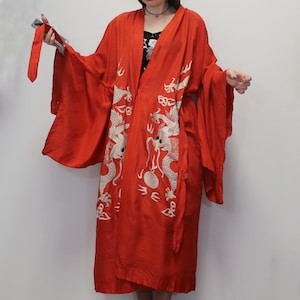 May include: A vibrant red kimono-style robe with long sleeves and a wrap-around design. The robe features intricate white embroidered dragon motifs on the front. The fabric appears to be a lightweight material, suitable for layering. The robe has a matching tie belt.