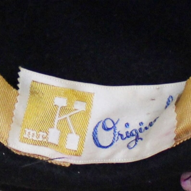 Puede incluir: Una etiqueta blanca con un rect&aacute;ngulo amarillo que contiene una "K" estilizada y el texto "K.S.T.R.". La etiqueta tambi&eacute;n tiene el texto "Origine" en escritura cursiva azul.