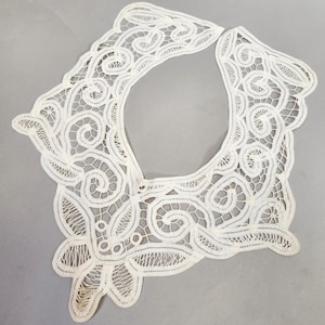 May include: A delicate, off-white lace collar with an intricate, openwork design. The collar features swirling patterns and scalloped edges, creating a vintage aesthetic. The lace is likely cotton or linen.