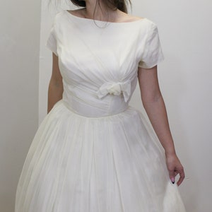 May include: A white vintage wedding dress with a fitted bodice and a full skirt. The bodice has a scoop neckline and short sleeves. A bow is located at the waist.