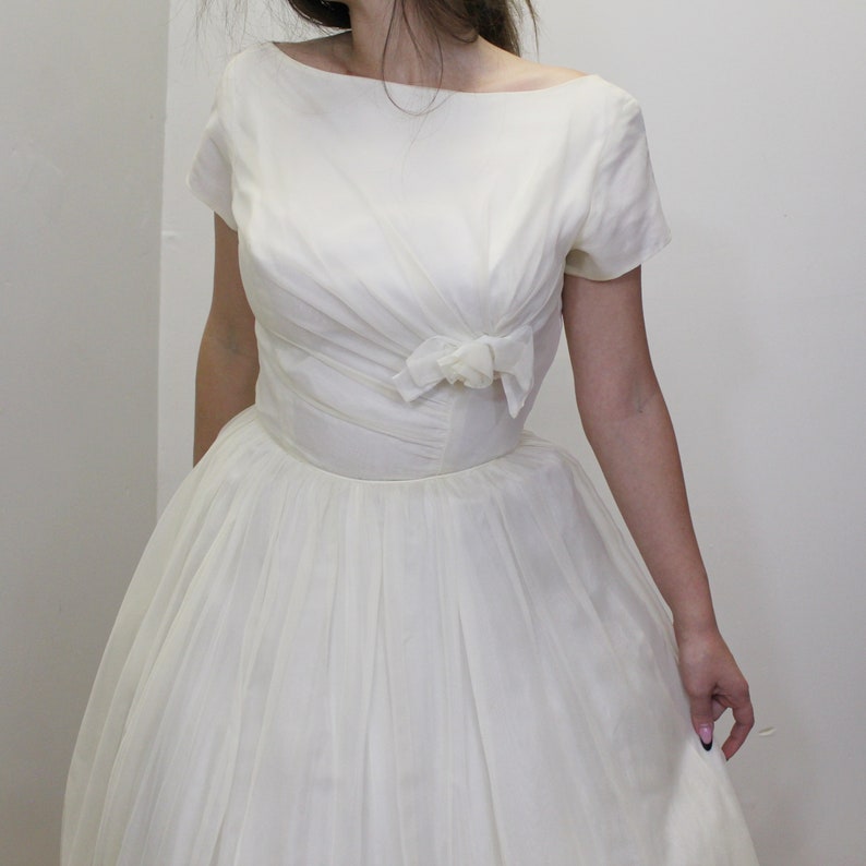May include: A white vintage wedding dress with a fitted bodice and a full skirt. The bodice has a scoop neckline and short sleeves. A bow is located at the waist.