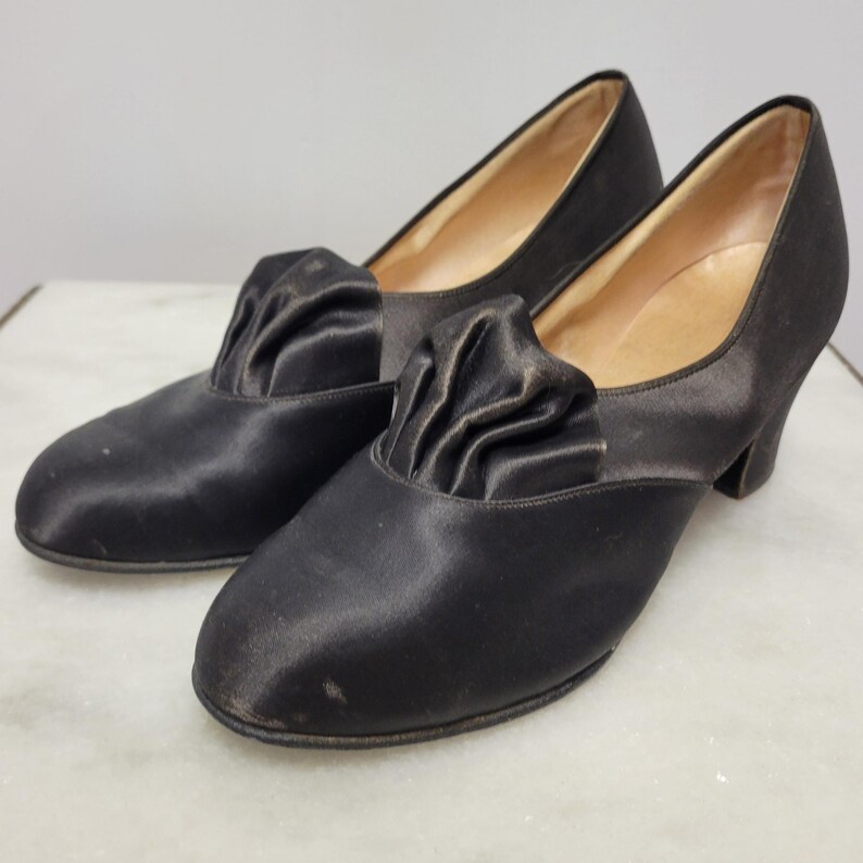 May include: A pair of black satin vintage shoes. Each shoe has a gathered fabric detail on the vamp and a small heel. The shoes have a classic, elegant design.