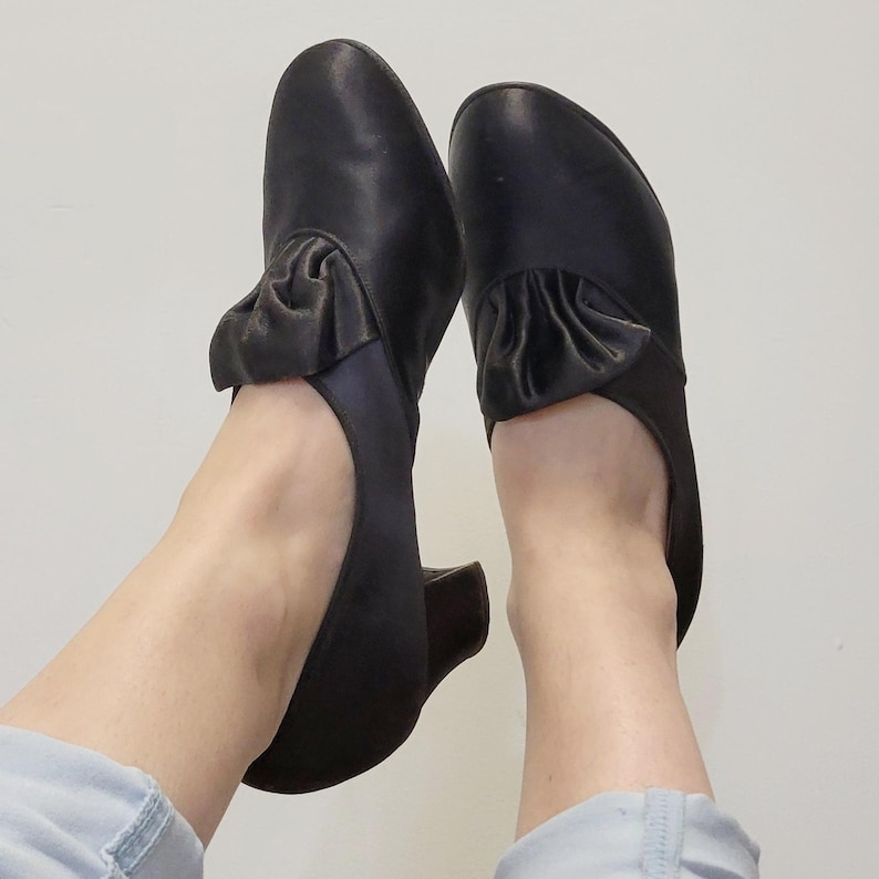 May include: A pair of black leather shoes with a low heel and a decorative ruffle detail on the top. The shoes have a classic design and are suitable for various occasions.