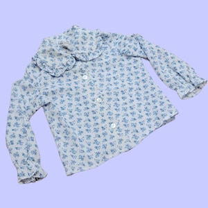 Pode incluir: Uma camisa branca de manga comprida com padrão floral azul e gola com babados.