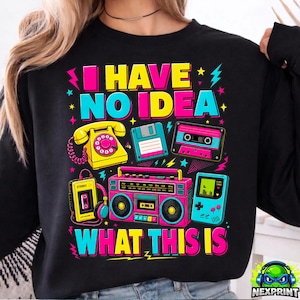Peut inclure: Sweat-shirt noir avec un motif graphique coloré représentant des appareils électroniques rétro tels qu'un téléphone, une cassette et un boombox. Le texte "I HAVE NO IDEA WHAT THIS IS" est affiché en couleurs vives.