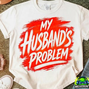 Könnte beinhalten: Weißes T-Shirt mit dem weißen Text "MY HUSBAND'S PROBLEM" auf einem roten Pinselstrich-Design. Ein lässiges Kleidungsstück.
