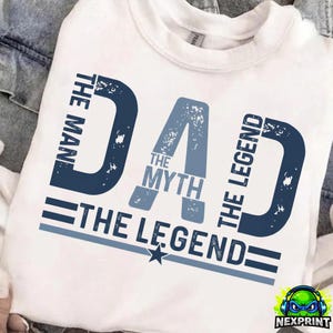 Könnte beinhalten: Weißes T-Shirt mit einem blauen Aufdruck mit der Aufschrift "DAD The Myth The Legend". Die Wörter "The Man" und "The Legend" sind vertikal auf beiden Seiten des Wortes "DAD" aufgedruckt. Ein Stern und zwei horizontale Linien befinden sich darunter.