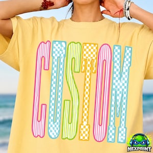 以下が含まれることがあります： 黄色いTシャツ。大きなカラフルな文字で「CUSTOM」と書かれています。文字はピンク、青、緑、黄色で縁取られ、ストライプとチェック柄があります。Tシャツは半袖で、クルーネックです。